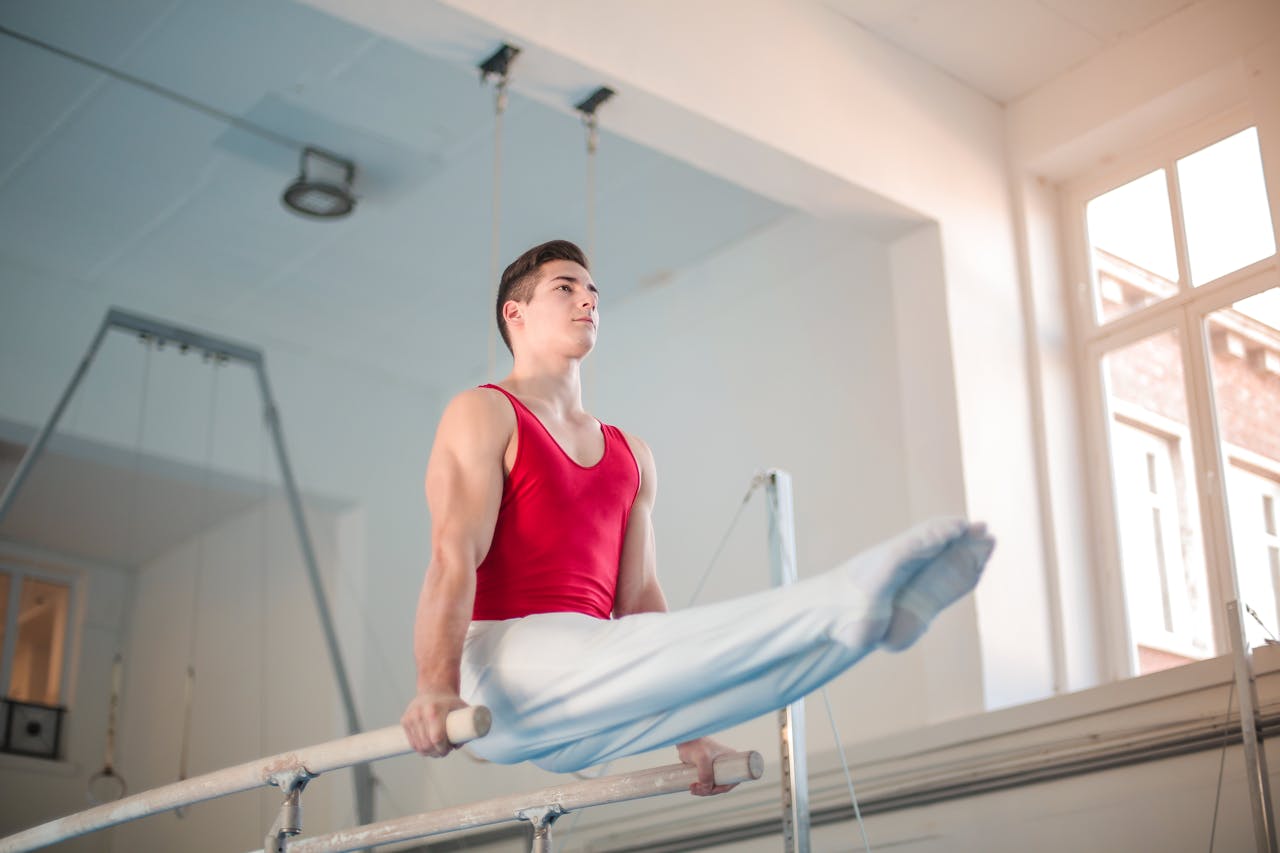 Young male gymnast in red top demonstrates strength on parallel bars indoors.